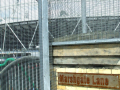 A rusted sign saying Marshgate Lane on a wall, beside fences with the Olympic stadium in the background