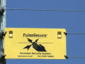 A yellow sign bearing a warning of a nearby electric fence, hanging from a grey fence